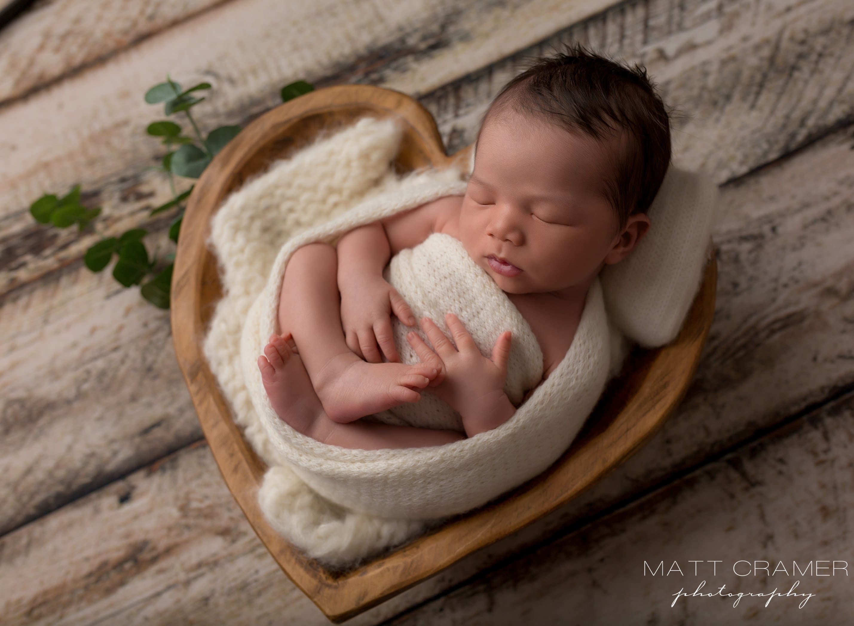 newborn baby boy in cream swaddle in wood heart prop during newborn photography photoshoot