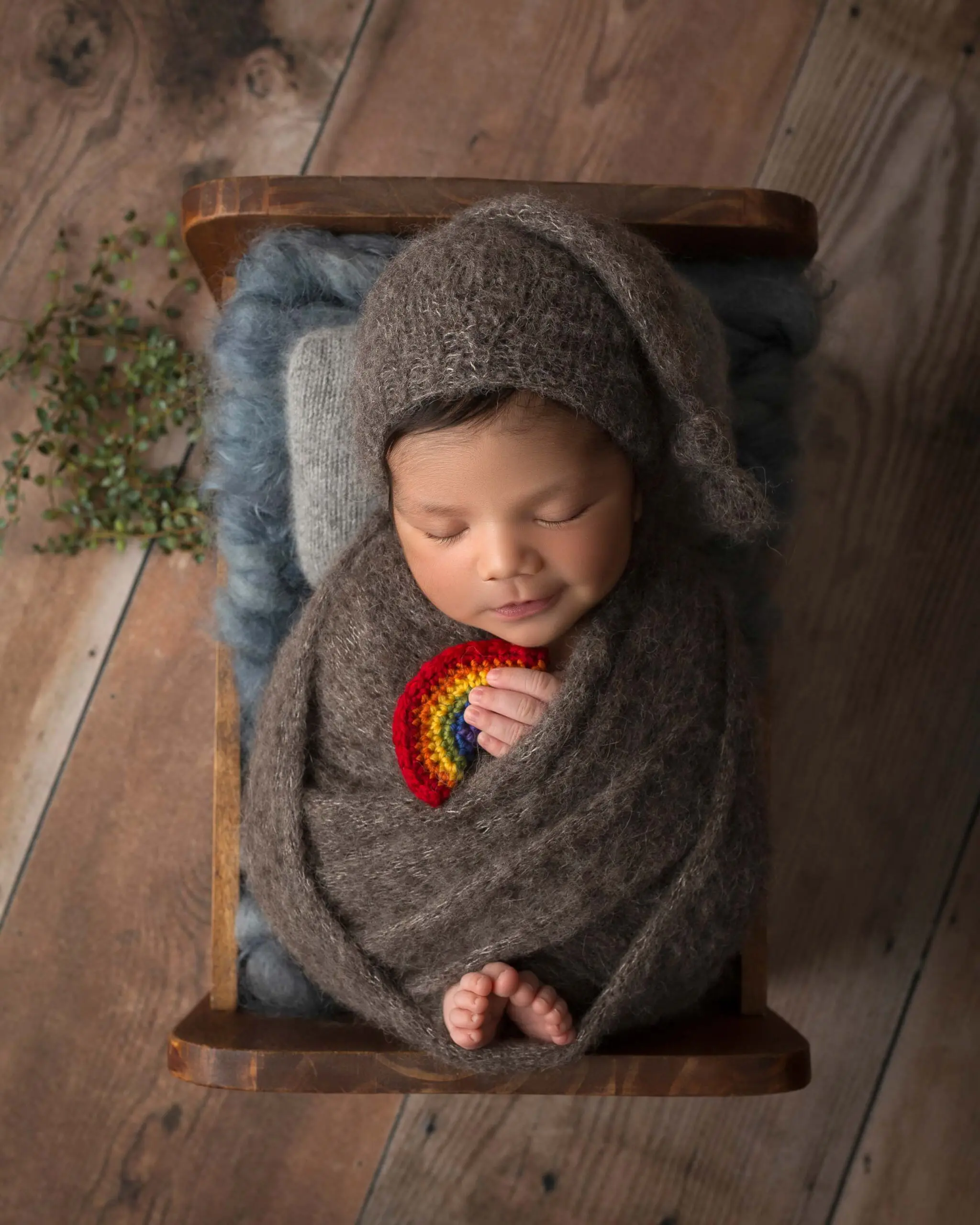 baby boy in dark grey swaddle wrap holding rainbow lovey