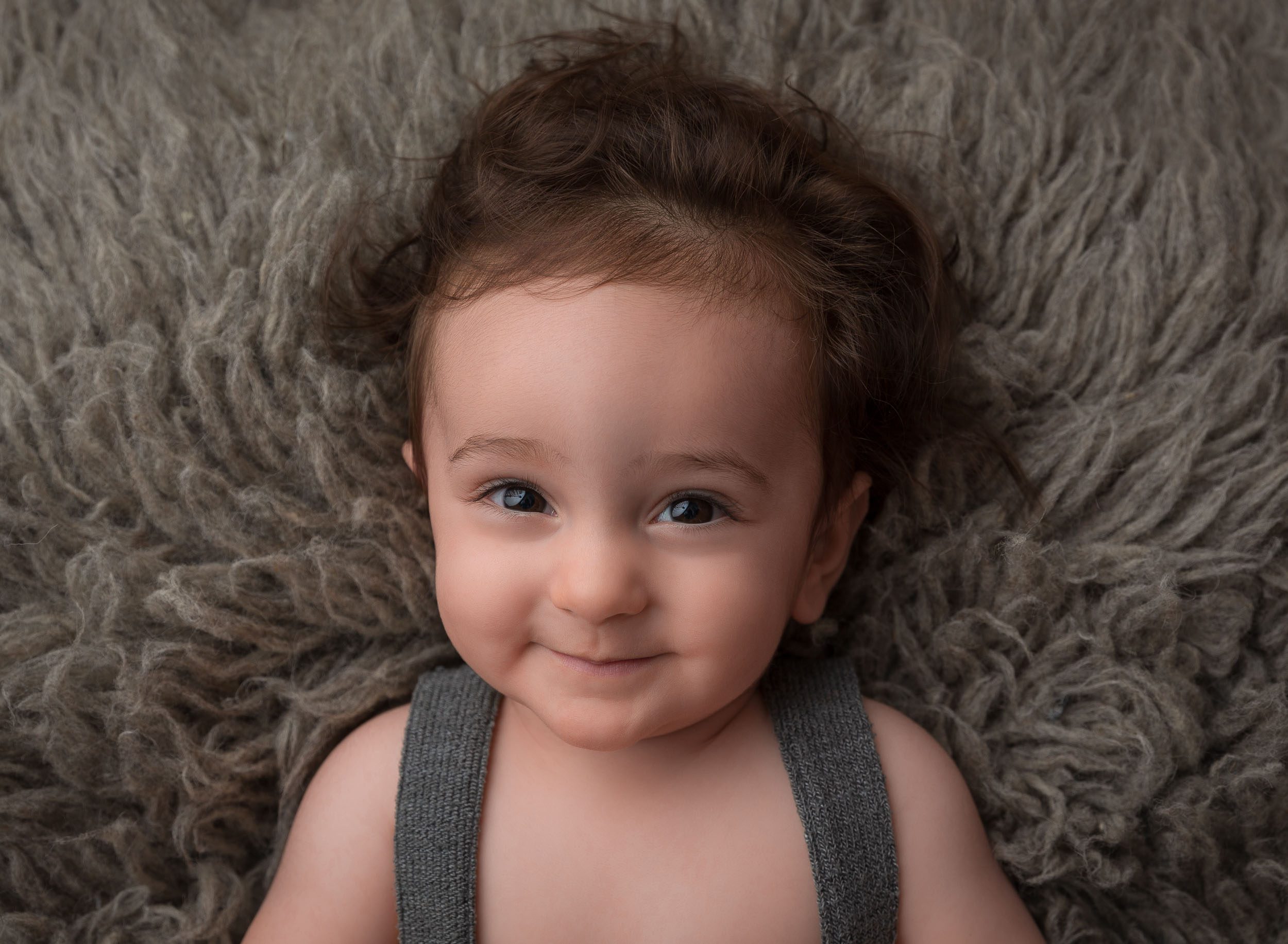 baby boy in overalls on grey rug looking up at camera