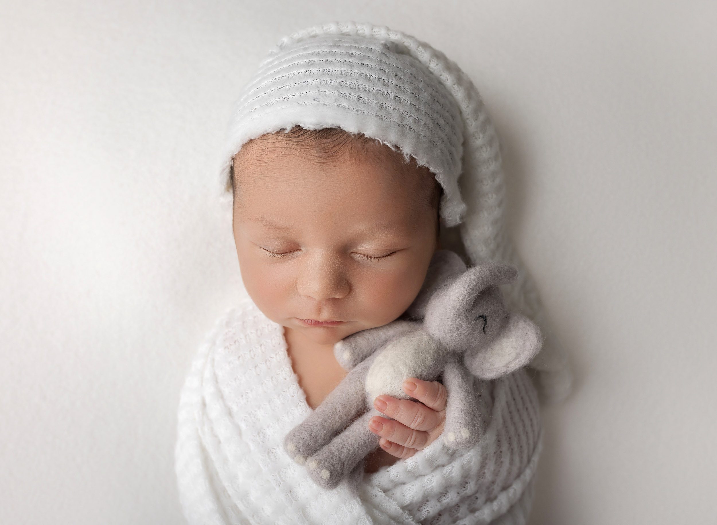 Newborn baby boy on white sleepy hat swaddled in matching whit fabric snuggling elepahant toy